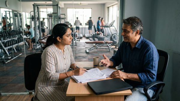 A gym owner and manager plan a software switch over printed papers at a desk overlooking the gym floor